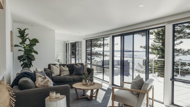 A modern living room with a sofa, armchair, and coffee table, large windows with a view of the sea, and a potted plant. - Sandy Point Beach House - On The Waterfront.  Mid Week Stay Special Is Now On. (Corlette)