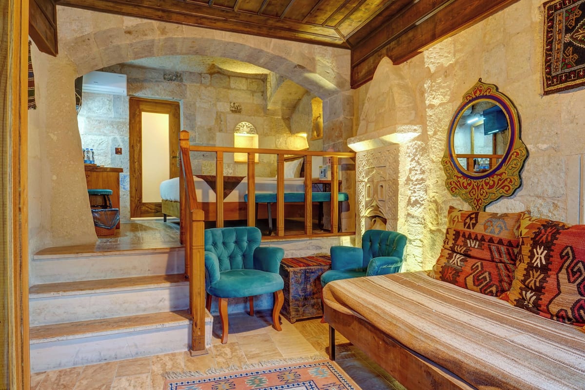 A room with a stone wall, a wooden staircase, a bed with patterned pillows, and a mirror with an ornate frame.