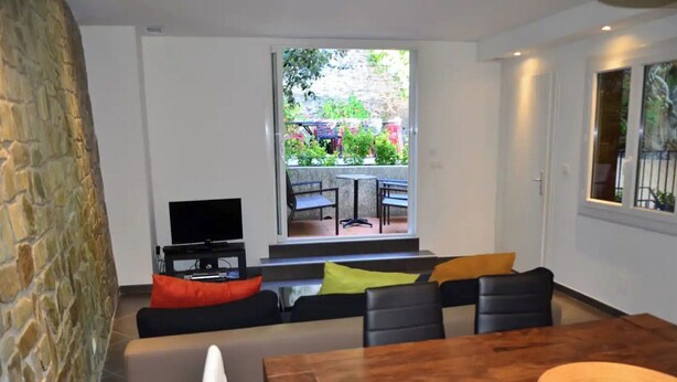 Una sala de estar moderna con una pared de piedra, un televisor, un sofá con cojines coloridos, una mesa de comedor de madera y vistas a un jardín a través de un ventanal grande.