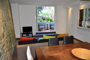 Uma sala de estar moderna com uma parede de pedra, uma televisão, um sofá com almofadas coloridas, uma mesa de jantar de madeira e vista para um jardim através de uma grande janela.