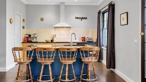 Een moderne keuken met een centraal eiland, vier houten krukken, een wit plafond en een bordje aan de muur met de tekst 'imagine'.