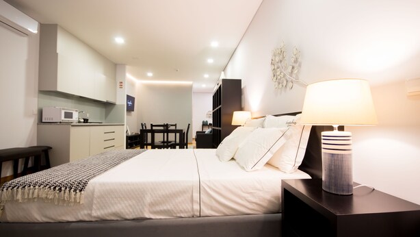 Quarto de hotel moderno com uma cama bem arrumada, uma mesa de cabeceira com abajur, uma pequena escrivaninha e uma cozinha compacta ao fundo.