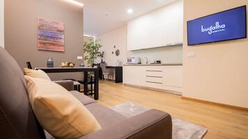 A modern living room with a sofa, a dining area with a table and chairs, a kitchen with white cabinets, and a flat-screen TV on the wall.