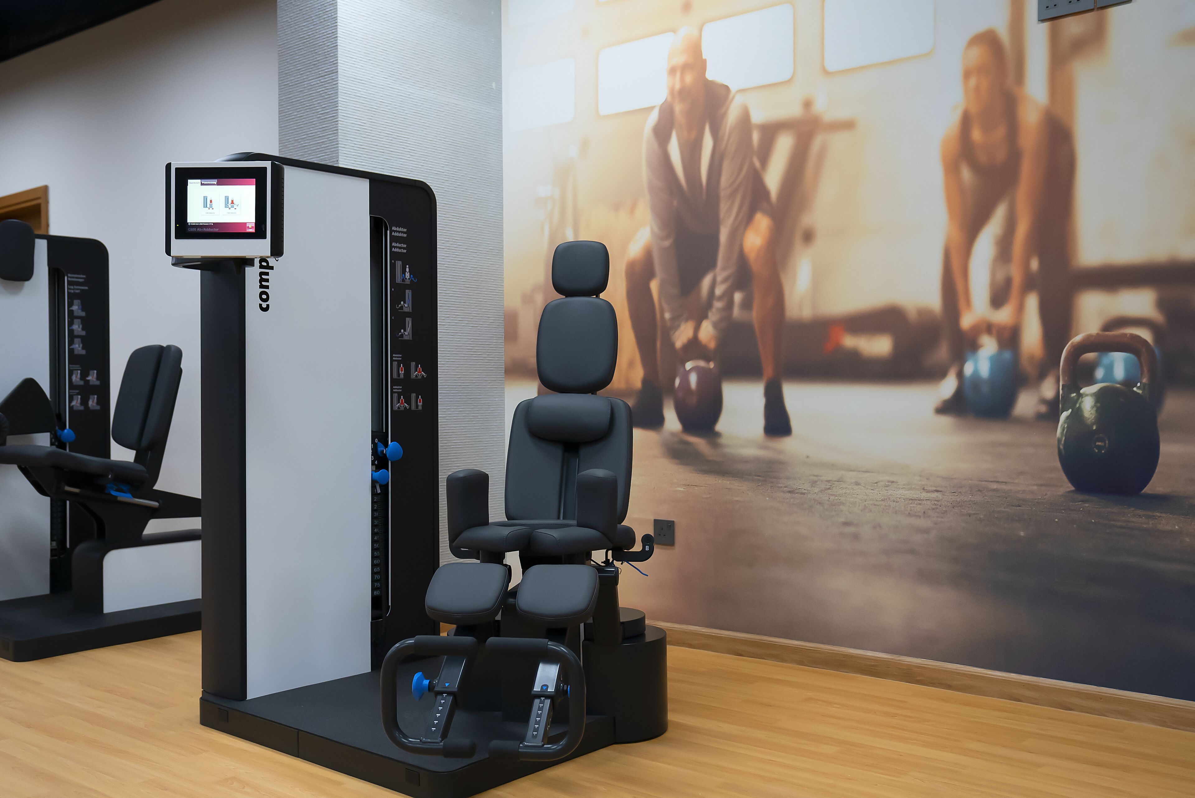 A gym with exercise equipment, a digital display, and a poster of people exercising.