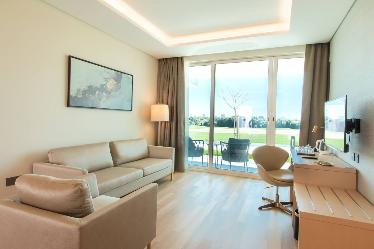 A modern hotel room with a sofa, a desk, a chair, a television, and a large window with a view of a golf course.