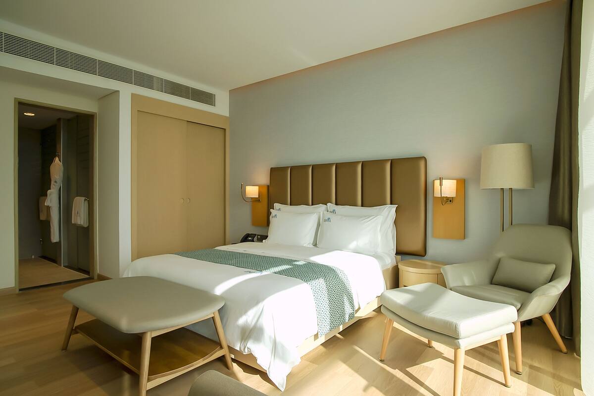 A modern hotel room with a large bed, a sitting area, a chair, and a wardrobe.