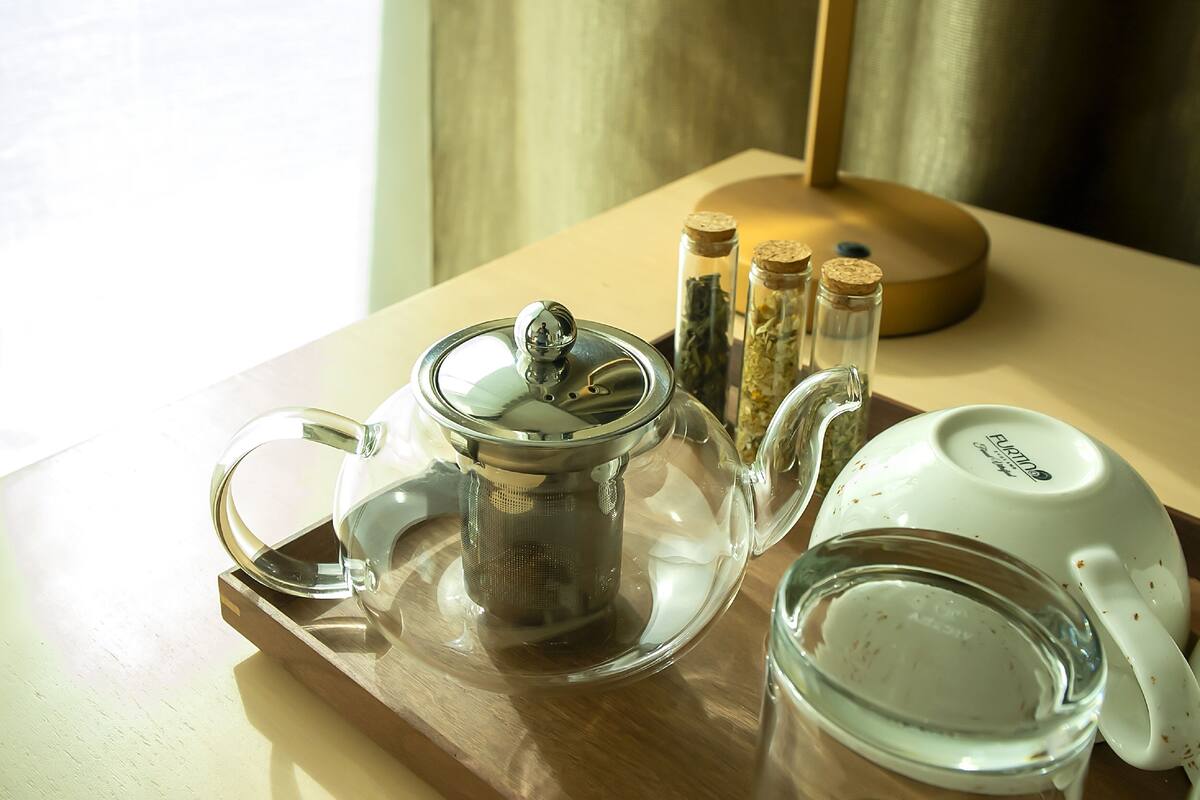 A wooden tray with a glass teapot, a glass cup, and two glass containers with cork stoppers.