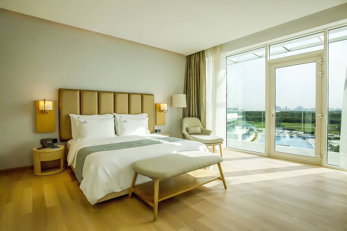 A modern hotel room with a large bed, a bench, a chair, and a view of a cityscape through large windows.