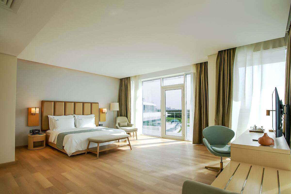 A hotel room with a bed, bedside tables, a desk with a chair, a TV, and large windows with curtains.