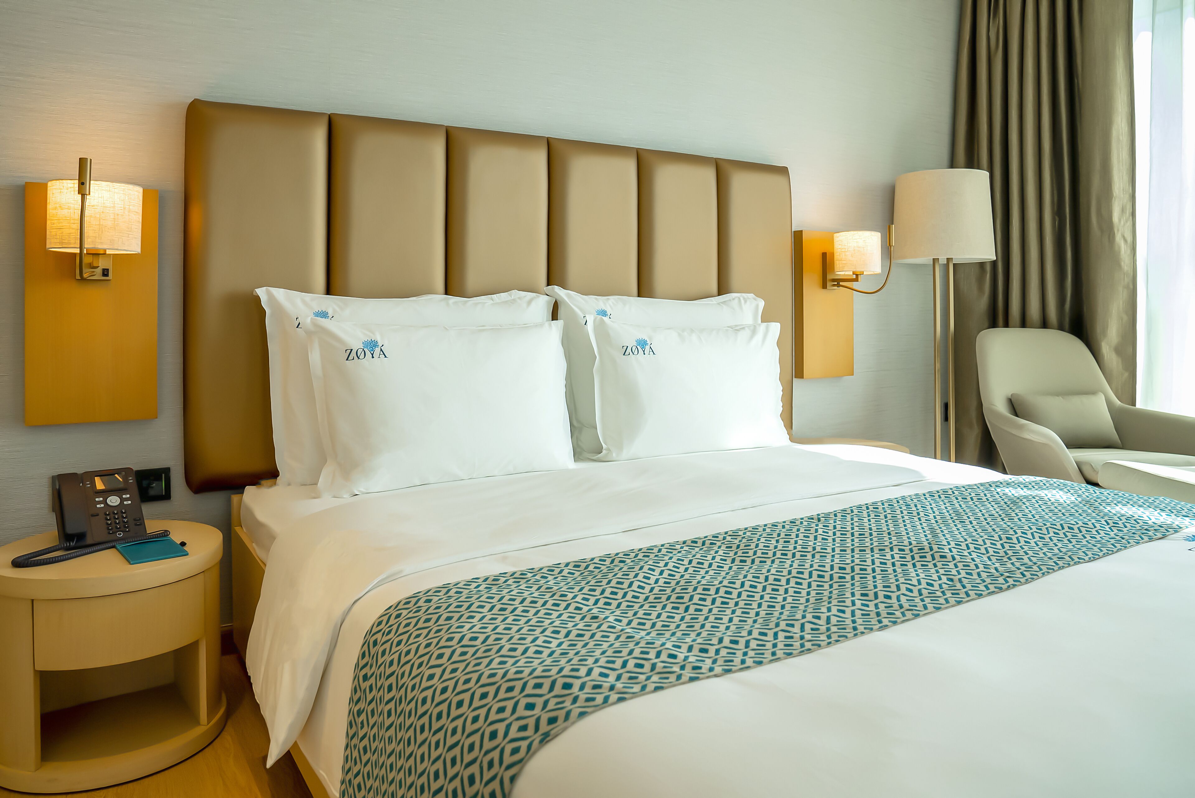 A hotel room with a large bed, two bedside lamps, a nightstand with a phone, and a chair.