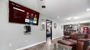 Un salon moderne avec un téléviseur fixé au mur qui affiche un écran d’accueil, une table basse en verre, un fauteuil inclinable en cuir et, au fond, un coin cuisine.