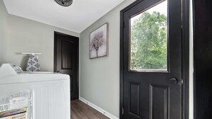 Un couloir avec une porte noire, un panier à linge blanc, un tableau encadré accroché au mur et, à travers la porte, une vue sur les arbres.