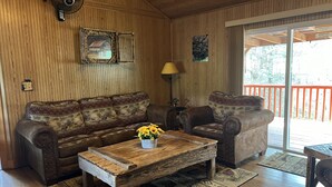 Un salón con paneles de madera, un sofá, sillones, una mesa de centro con un jarrón de girasoles y vistas a una terraza roja a través de un ventanal.