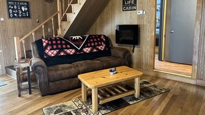Interior de una cabaña con una escalera de madera, un sofá de cuero marrón, una mesa de centro de madera y un televisor montado en la pared.