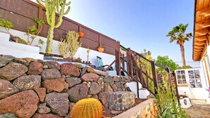 A stone retaining wall with various cacti and plants, a wooden pergola, and a white building with a window. - Vacation apartment CASA LA ISLA, Tarajalejo, fiber optic internet (Tarajalejo)