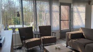 Ein modernes Wohnzimmer mit einer Couch, zwei Sesseln, einem kleinen Tisch und Blick auf einen Garten durch große Fenster.