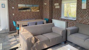 Ein gemütliches Zimmer mit Backsteinwand, einem Holzesstisch mit Stühlen, einem grauen Sofa und einem Fenster mit Jalousien.