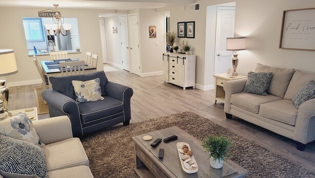 A living room with a dark blue sofa, a beige sofa, a wooden coffee table, and a beige couch. There is a ceiling fan, a chandelier, and a dining area in the background. - Ocean view flat, poolside ( ground floor). Two  bedrooms, two baths. (St. Augustine)
