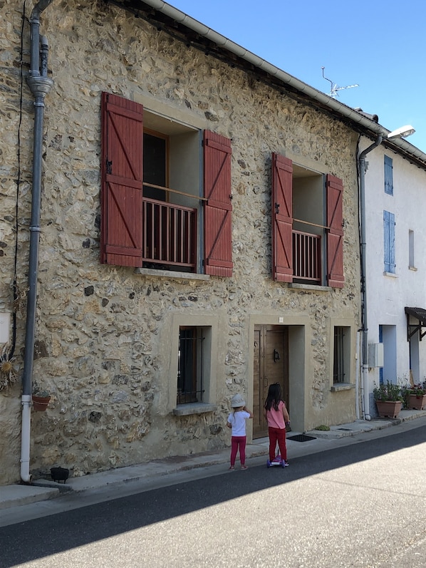 A stone building with two windows featuring red shutters, a small balcony, and a door.