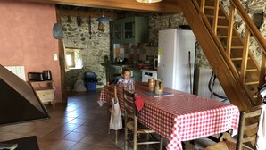 Uma cozinha rústica com parede de pedra, vigas de madeira, mesa coberta por um tecido xadrez e uma criança à mesa.