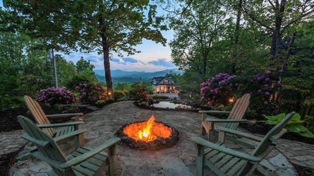 A stone patio with a fire pit, surrounded by Adirondack chairs, and a view of a house in the background. - High View Heights stunning views and complete privacy (Bryson City)