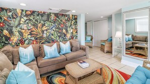 A living room with a brown sofa, blue cushions, a wicker coffee table, a colorful rug, a large mirror, and a floral wall mural. - Oceanfront Tybee Cottage | Private Hot tub & Beach Access (Tybee Island)