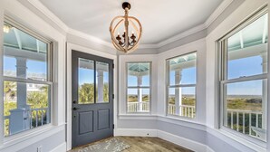 Una habitación con una puerta gris oscuro, ventanas grandes con marcos blancos, suelo de madera y un candelabro.