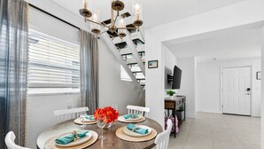 A dining area with a round table set for four, white chairs, a chandelier, and a staircase leading to another level. - Frenchy’s Cottage 480 in Clearwater Beach (Clearwater Beach)