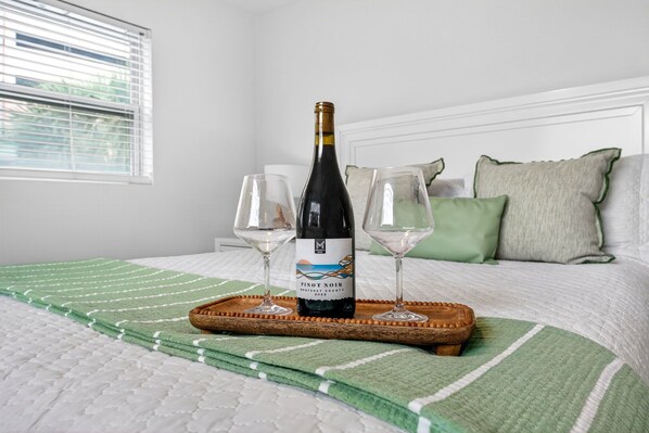 Un lit recouvert d’une couverture à rayures vertes et blanches, une bouteille de vin Pinot Noir et deux verres à vin vides sur un plateau en bois.