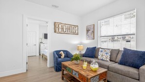 A modern living room with a wooden coffee table, a grey sofa, and a blue armchair. A 'BEACHHOUSE' sign is on the wall, and a window with blinds is visible.