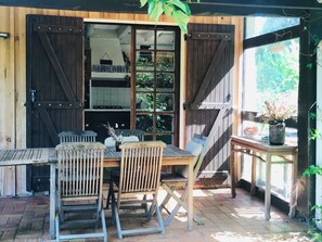 Una terraza de madera con una mesa y sillas, una planta colgante y vistas del interior a través de una puerta de vidrio.