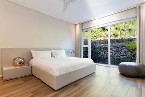 A modern bedroom with a large bed, a bedside table, a lamp, and a view of a garden through a sliding glass door. - Thabisa Penthouse Supreme Views & Exquisite Design (Cape Town)