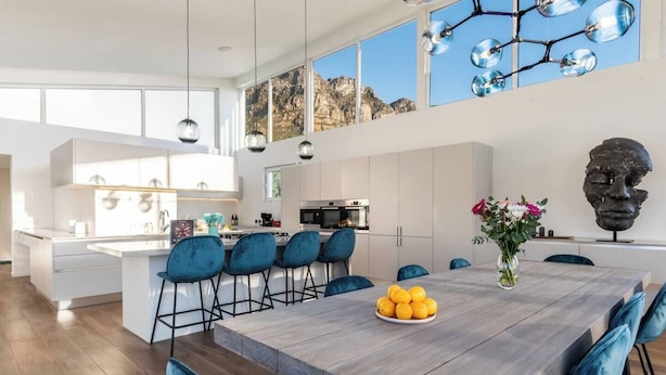 A modern kitchen with a central island, bar stools, and a dining table with a fruit bowl.