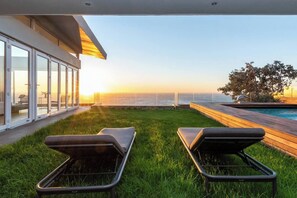 Ein moderner Außenbereich zum Entspannen mit zwei Liegestühlen, einem Pool und Blick auf das Meer bei Sonnenuntergang.