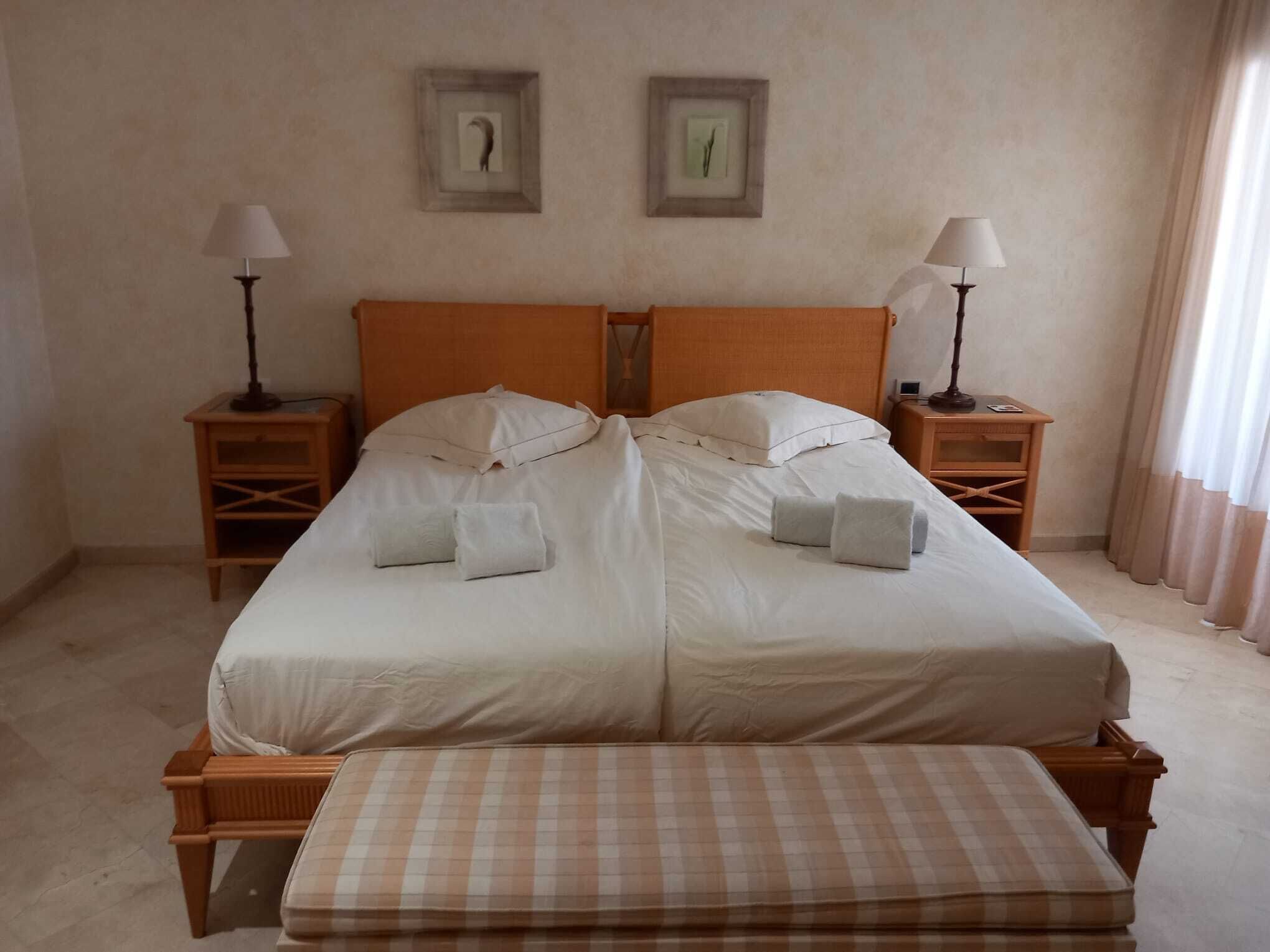 Una cama doble con sábanas blancas, dos mesitas de noche con lámparas, y un banco a cuadros al pie de la cama.