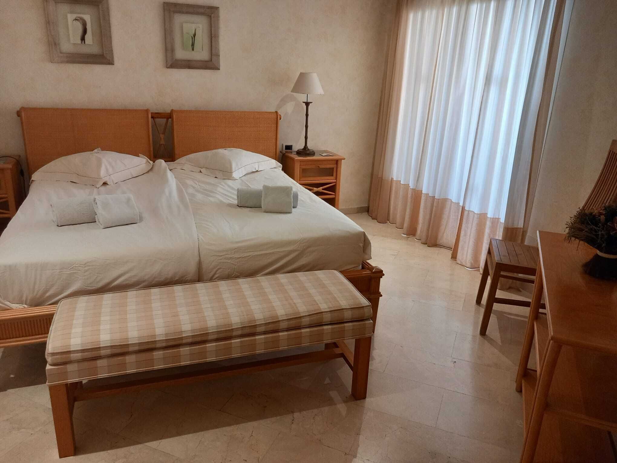 A neatly arranged bedroom with a large bed, a bedside table with a lamp, a bench, and a small table with a plant.