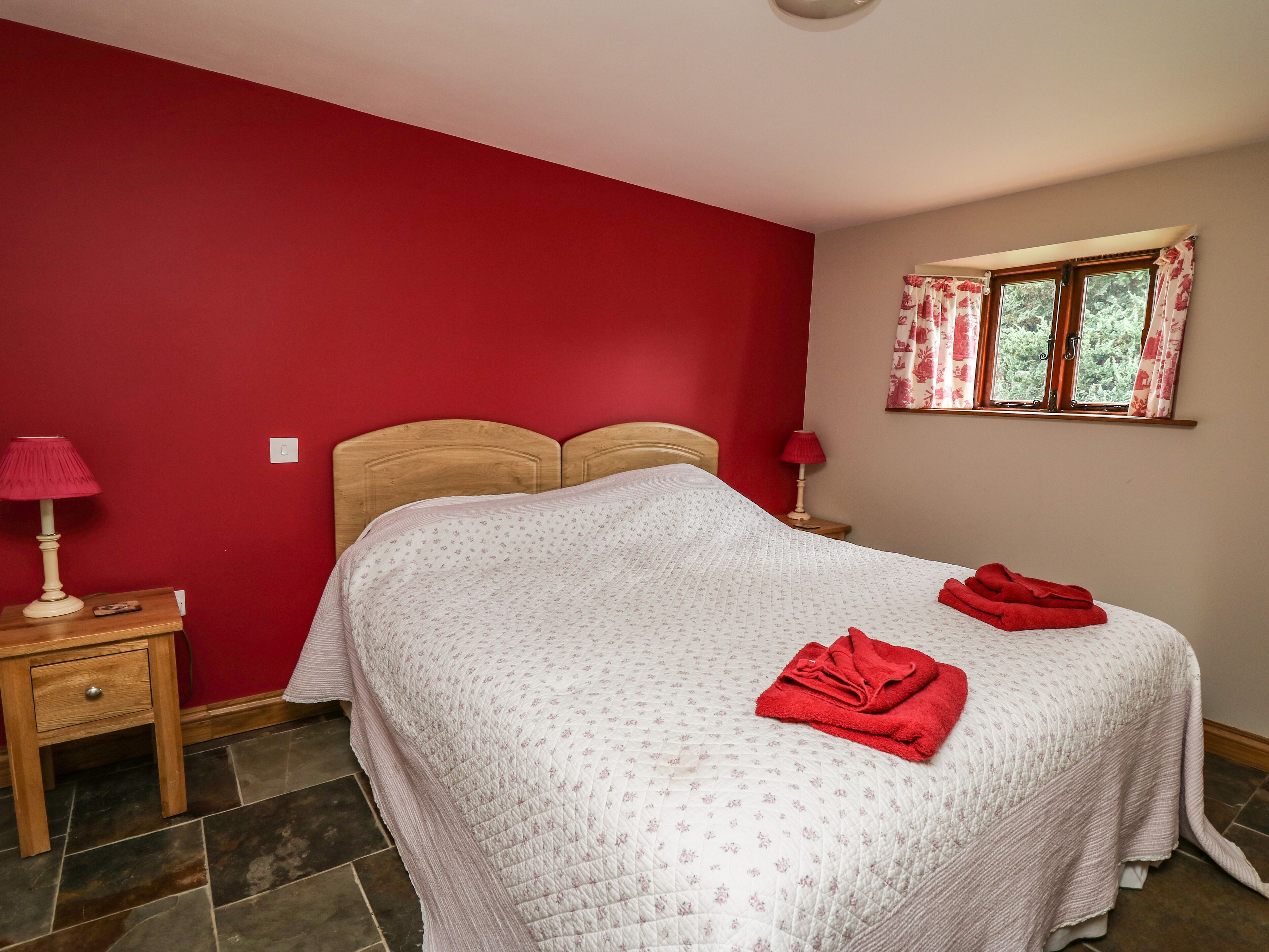 Une chambre à coucher avec un lit, une table de chevet, une lampe, une fenêtre avec des rideaux et un mur rouge.
