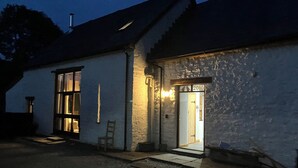 Un bâtiment blanc avec une façade en pierre, une porte en bois surmontée d’une enseigne, et un petit porche avec une chaise.