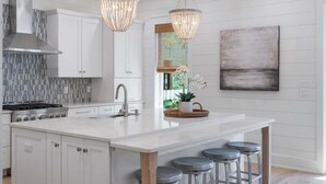 Une cuisine moderne avec des meubles blancs, un grand îlot central doté de tabourets, une cuisinière à gaz et deux luminaires suspendus.