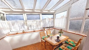 Un solárium con suelo de baldosas, una mesa de madera con sillas, y vistas a través de amplios ventanales.