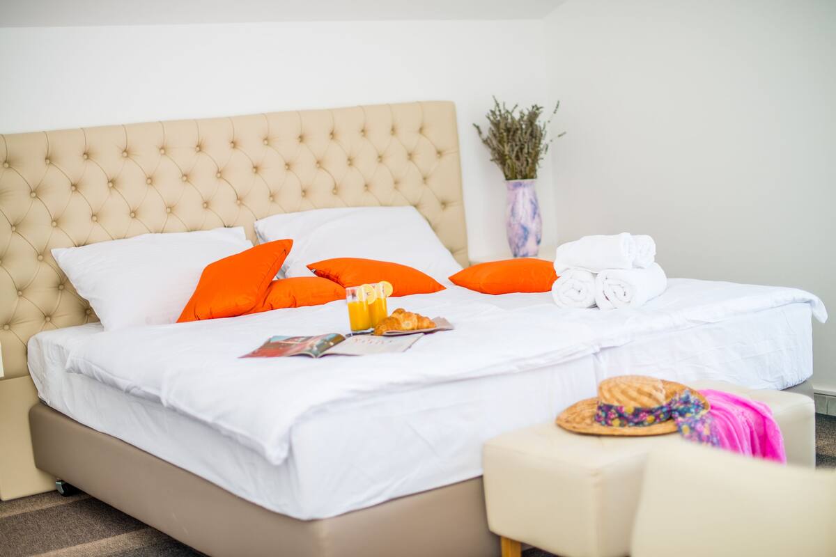 A neatly made bed with white and orange pillows, a white duvet, and a small table with a hat and a magazine.