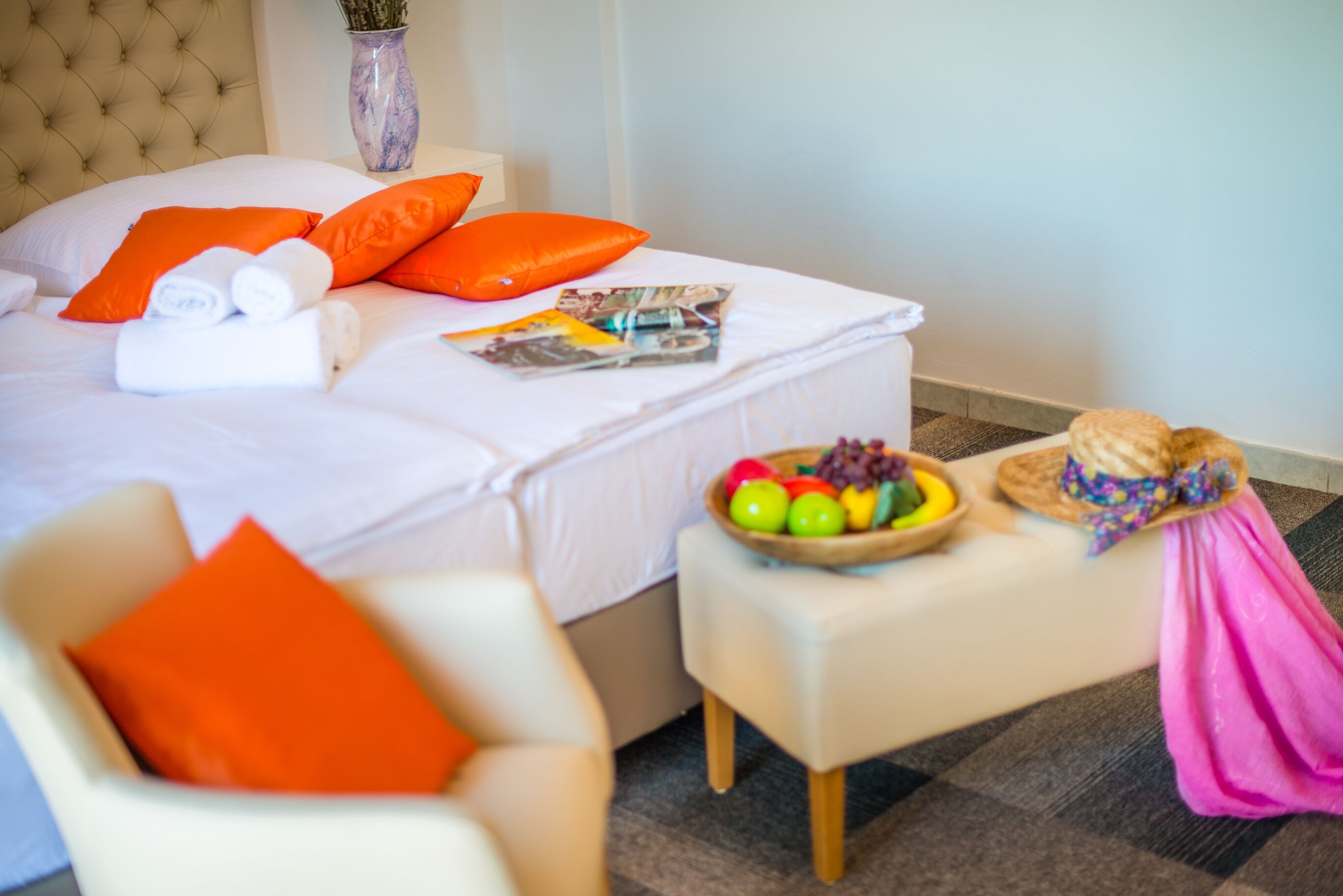 A neatly made bed with orange pillows, a small table with a fruit bowl, and a chair with an orange pillow.