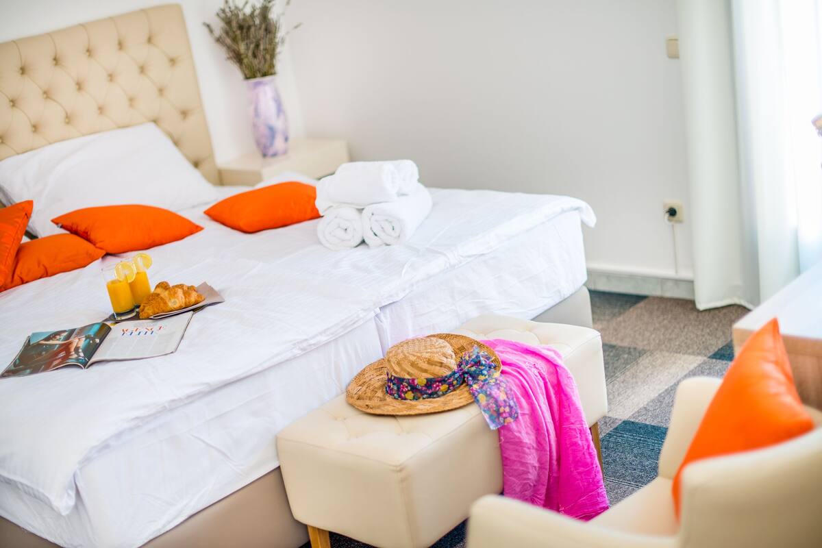 A neatly made bed with white linens, orange pillows, and a rolled towel. There is a magazine, a straw hat, and a pink towel on the bed.