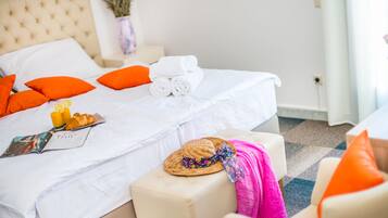 Una cama bien hecha con sábanas blancas, almohadas naranjas y una toalla enrollada. Hay una revista, un sombrero de paja y una toalla rosa sobre la cama.