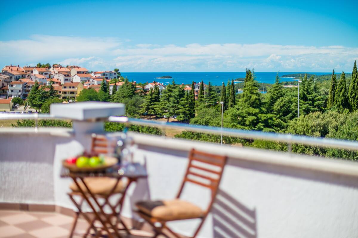 A balcony with a table and chairs, overlooking a coastal town with houses and greenery.