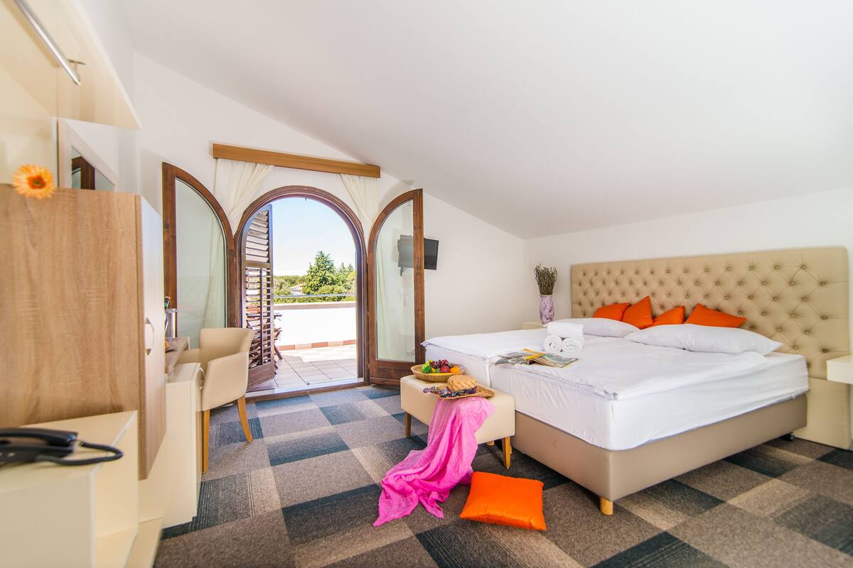 A hotel room with a bed, a desk, a chair, a small table with a basket of fruit, and a view of the outdoors.