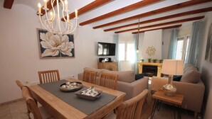 Un comedor con mesa de madera, sillas y una araña. Se observa un sofá y un televisor al fondo.