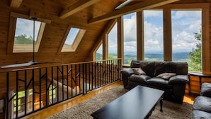 A wooden interior with a black leather sofa, a small table, and a view of the outdoors through large windows. - Mountain View Sanctuary | Pet-Friendly Weaverville Log Cabin | Hot Tub & Long-Range Views (Weaverville)