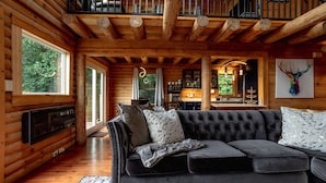A log cabin interior with a dark grey sofa, wooden beams, a fireplace, and a view of the outdoors. - Mountain View Sanctuary | Pet-Friendly Weaverville Log Cabin | Hot Tub & Long-Range Views (Weaverville)