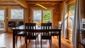 A wooden cabin interior with a dining table and chairs, large windows, and a view of the outdoors. - Mountain View Sanctuary | Pet-Friendly Weaverville Log Cabin | Hot Tub & Long-Range Views (Weaverville)